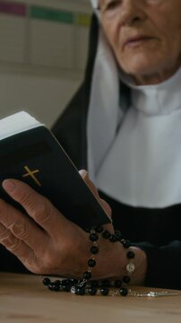 Vertical of senior Caucasian nun in veil sitting at table and reading Bible out loud teaching in convent or in religious school