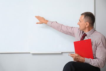 Male teacher with clipboard sitting on chair in classroom and pointing at flipboard