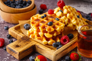 Wooden board of tasty waffles with berries and maple syrup on grey background
