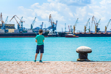 Boy fishing on sunny pier with industrial port in background, standing near turquoise water. Concept of fishing, childhood moments, peaceful outdoor leisure