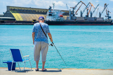 Senior man fishing near port waterfront, holding rod beside blue chair and bucket under sunlight. Concept of fishing, peaceful routine, retirement lifestyle by seaside