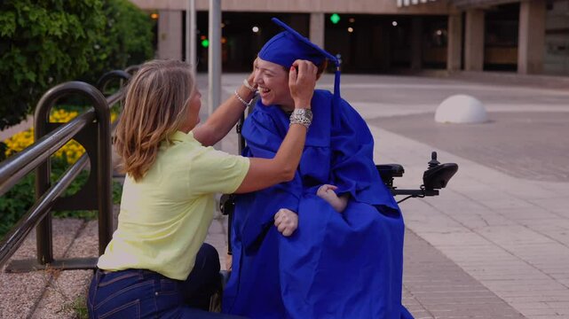 Happy mother adjusts her disabled daughter's mortarboard, who is wearing a blue graduation gown and sitting in a wheelchair, in front of a modern building