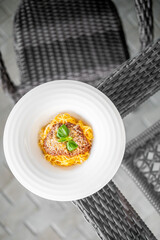 Plate of spaghetti with a grilled meat patty and fresh basil garnish served on a wicker table outdoors, evoking a cozy, rustic dining atmosphere with natural textures and soft light