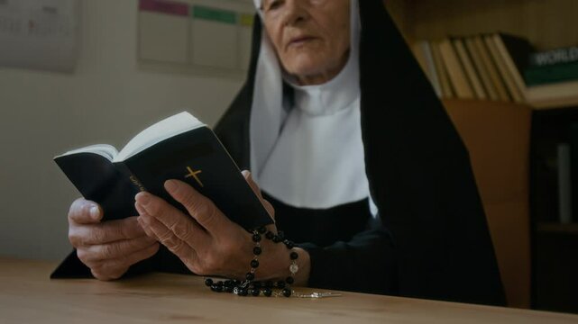 Low angle shot of senior Caucasian nun in veil sitting at table and reading Bible out loud during Bible class in convent or in religious school