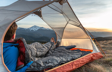 Man lying in tent with sleeping bag, watching mountain sunset view, camping in the wilderness,...