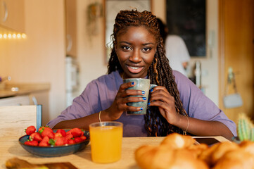 Smiling young woman drinking a hot beverage at home, enjoying a healthy breakfast with fresh fruit,...