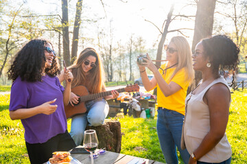 Four joyful friends enjoying picnic, taking photos and playing guitar in park