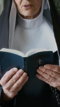 Vertical cropped shot of focused senior Caucasian nun in veil reading Bible out loud indoors