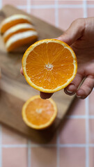 Close-up of a hand holding a halved orange. In the background, sliced oranges rest on a wooden board. Captured in warm natural light for a fresh and vibrant pink aesthetic.