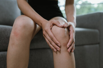 A woman sits indoors holding her painful knee, showing signs of joint inflammation or injury. Red...