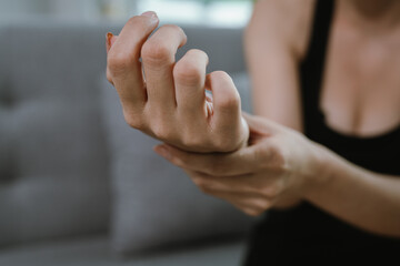 A woman sitting indoors holds her painful wrist with both hands, showing signs of discomfort,...
