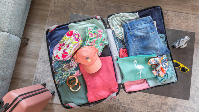 View from above of open suitcase placed on coffee table with colorful summer clothes and accessories, getting ready for vacation, next to vintage photographic camera and yellow sunglasses