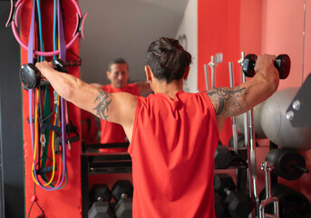 Strong sportsman wearing red sleeveless shirt performing lateral raise exercise with dumbbells in a...