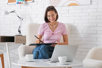 Young female graphic designer working with graphics tablet on armchair in office