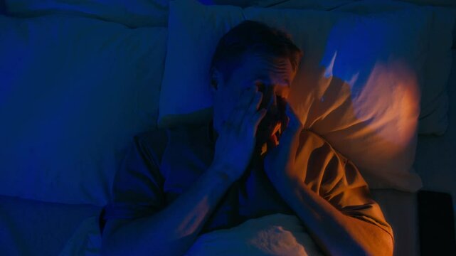 High angle shot of Caucasian man rubbing eyes and yawning while lying in bed and trying to get up