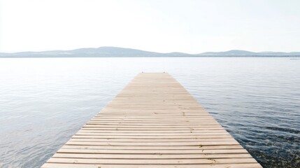 Obraz premium Tranquil wooden pier extending into calm water with weathered planks, patches of greenery, clear pebbly edges, deep blue expanse, and distant hazy mountains blending with pale sky.