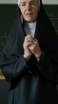 Vertical zoom out shot of elderly Caucasian nun in veil praying before starting religious class in Christian school
