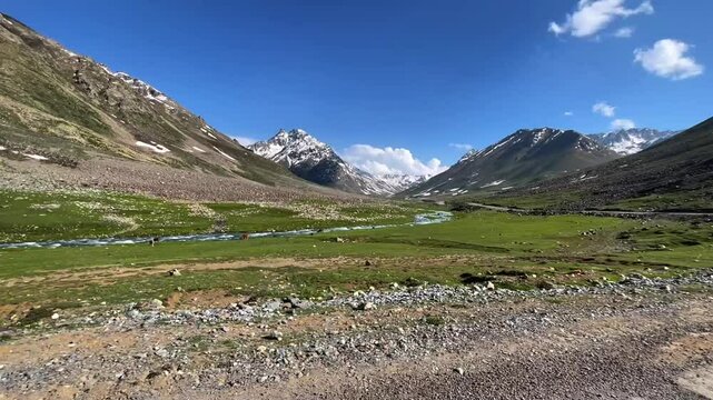 Cinematic video of Gitti Dass near Babusar Top, showing lush green meadows, flowing water from Lulusar Lake, and distant snow-covered Naran mountains. A peaceful natural landscape.

