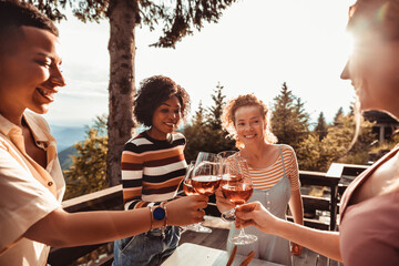 Young and diverse group of female friends enjoying a glass of wine and dinner on a balcony in a cabin house with a scenic view of the mountain and forest below