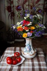 Field flowers in vintage vase and tomato branch on plate standing indoors on the checkered tablecloth. Colorful wild flowers bunch: yellow heliopsis, consolida, lychnis chalcedonica, cornflower, tansy