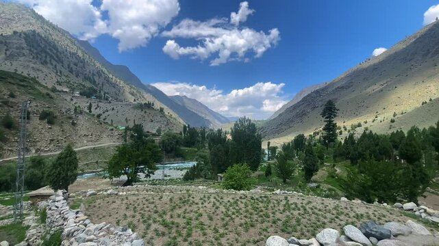 A peaceful valley view featuring crystal-clear blue river flowing between mountains, with a green village in sight. Blue sky with fluffy clouds enhances the serene atmosphere of this beautiful natural