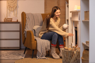 Young woman with cocoa sitting on armchair near fireplace at home in evening