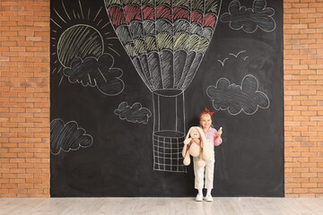 Cute little girl with chalk piece and toy near drawn hot air balloon on black wall