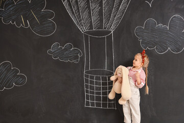 Cute little girl with chalk piece and toy near drawn hot air balloon on black wall