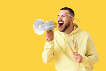 Young man with paint brushes shouting into megaphone on yellow background. Artist as Outlaw Day