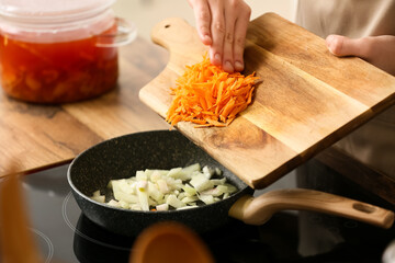 Young woman adding grated carrot into frying pan while cooking delicious borscht in kitchen, closeup