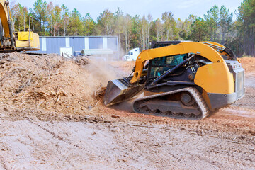 Heavy bulldozers machinery clears soil, debris at construction site surrounded by trees on a sunny...