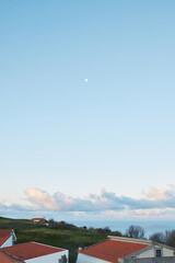 Moon in Blue Sky over Coastal Landscape with Houses and Red Roofs