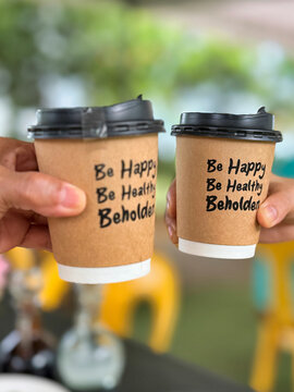 hands hold up takeaway coffee cups with black lids and motivational text that reads, &ldquo;Be Happy, Be Healthy, Behold,&rdquo; in an outdoor setting with blurred green trees and yellow chairs in the background.