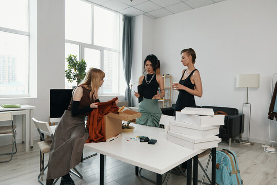 Three young adult women coworkers socializing and working together in a bright workplace, unpacking boxes and discussing business in a modern office environment.