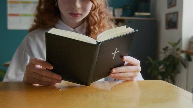 Cropped shot of beautiful red-haired teen Caucasian girl in white shirt reading Bible sitting at single desk in classroom