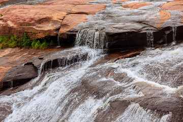 Clear waterfall flows steadily down rugged rock face, surrounded by lush greenery under bright...