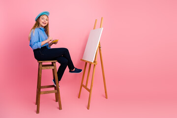 Young girl artist in blue attire smiling next to canvas with pink background radiating creativity and fun