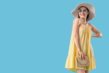 Stylish young woman in summer hat with sunglasses and bag on blue background