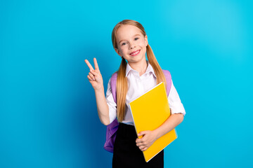 Smiling schoolgirl in uniform holding a notebook and making a peace gesture against a vibrant blue...