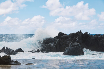 Powerful ocean wave crashes against dark volcanic rocks creating dramatic splash and foam by a...