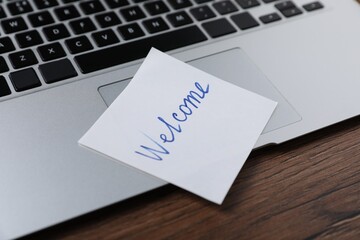 Paper note with word Welcome and laptop on wooden table, closeup