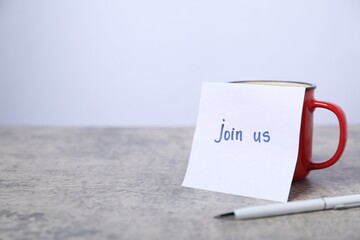 Sticky note with phrase Join Us, coffee and pen on grey table, closeup. Space for text