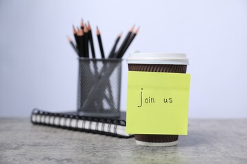 Sticky note with phrase Join Us, coffee, notebook and pencils on grey table