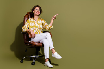 Young businesswoman in elegant blouse sits confidently in chair against khaki backdrop pointing...