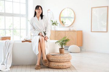 Young woman in soft bathrobe sitting on bathtub at home