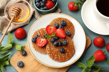 Tasty Dutch waffles (stroopwafels) with honey, berries and mint on light blue wooden table, flat lay