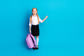 Cute schoolgirl pointing with hand on blue background, wearing uniform and holding backpack, ready...