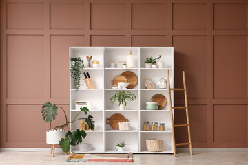 Shelf unit with kitchen utensils, plants and ladder near brown wall in room