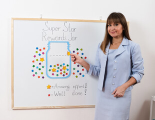A smiling female teacher stands beside a Super Star Rewards Jar board, placing colorful stars to...