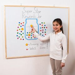 A cheerful schoolgirl places a bright star on a reward chart labeled “Super Star Rewards Jar.”...